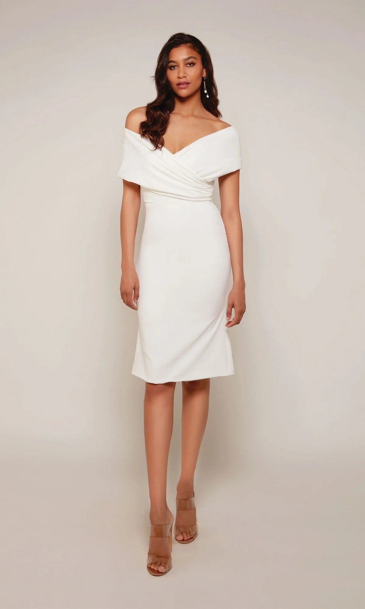 A woman stands against a plain background wearing an ivory Knee Length White Party Dress: Alyce 70023 and nude open-toe heels.